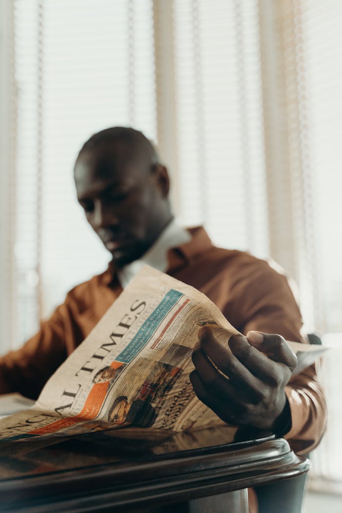 Man in brown shirt reading a newspaper by a window, relaxed and thoughtful setting.