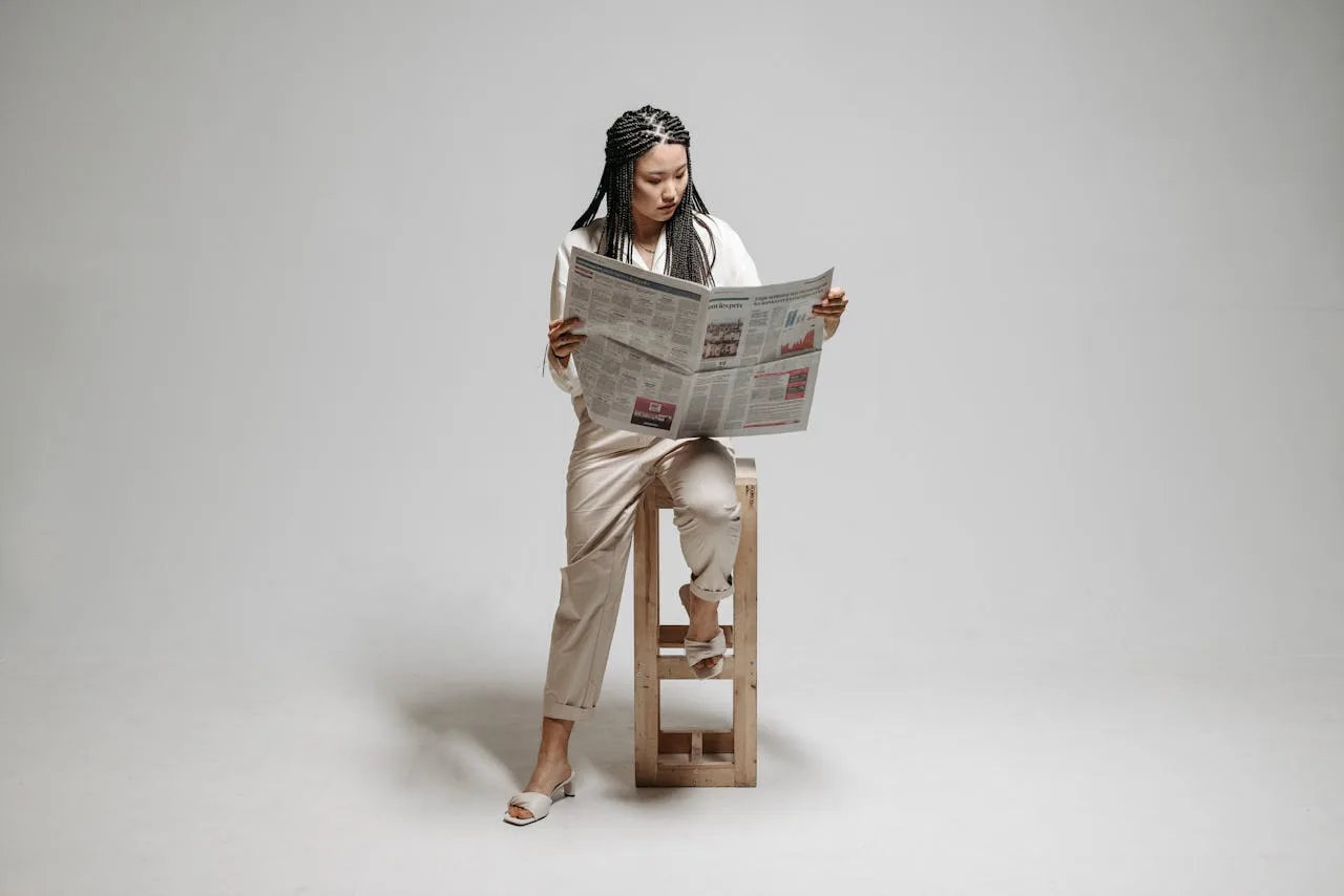 A woman sits on a stool with a braided hairstyle, reading a newspaper in a minimalistic studio.
